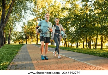Full length image of beautiful sporty healthy active cheerful middle age couple going to exercise outdoors in park holding mats for yoga, pilates, gym. Sports healthy lifestyle. Loving older couple.
