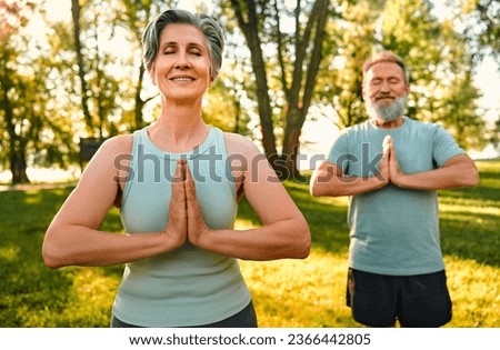 Yoga on retirement. Front view of mature man and woman dressed in black and blue outfits keeping hands in namaste gesture and meditating outdoors. Smiling couple closing eyes and doing yoga exercises. Royalty-Free Stock Photo #2366442805