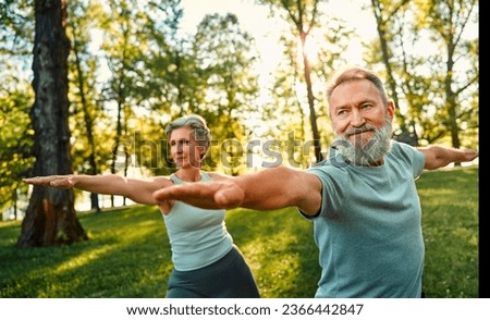 Yoga on retirement. Aged couple training outdoors at summer time and performing warrior asana exercise. Concentrated people standing in green forest with outstretched arms and practicing yoga poses.