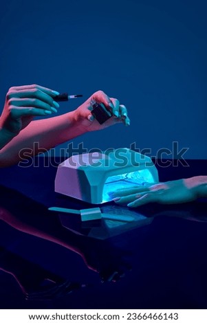 Manicurist using nail gel polish. Female client's hand into into nail uv lamp , drying gel against blue background in neon light. Concept of hand care, cosmetics and cosmetology, spa, natural beauty Royalty-Free Stock Photo #2366466143