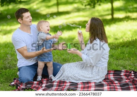A young cheerful family with a baby is having a picnic in the park,blowing soap bubbles. Summer time concept.Family day Royalty-Free Stock Photo #2366478037