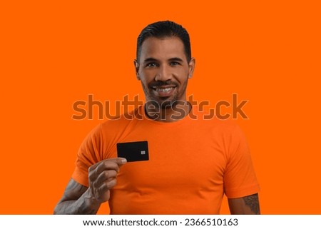 Man with a black credit card in his hand, wearing an orange T-shirt, looks at the camera and smiles, orange background