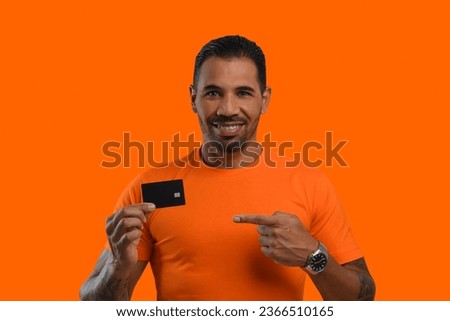 Man with a black credit card in his hand, points to the card, wears an orange T-shirt, looks at the camera and smiles, orange background