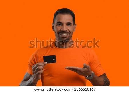 Man with a black credit card in his hand, points to the card, wears an orange T-shirt, looks at the camera and smiles, orange background