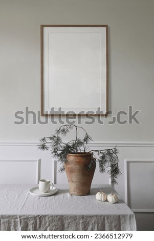 Elegant interior, Christmas table setting decor. Winter art composition. Blank wooden picture frame mockup hanging on beige wall. Pine tree branches in vase. Cup of coffee, white pumpkins. Vertical.