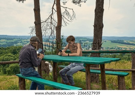 Grandfather and grandson are sitting at the table in the bosom of nature. An old man and a teenager look at smartphones. Family. Interests of generations. Nowadays communication. Royalty-Free Stock Photo #2366527939