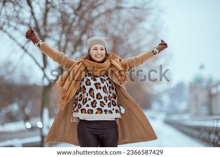smiling stylish woman in brown hat and scarf in camel coat with gloves and raised arms rejoicing outside in the city in winter. Royalty-Free Stock Photo #2366587429
