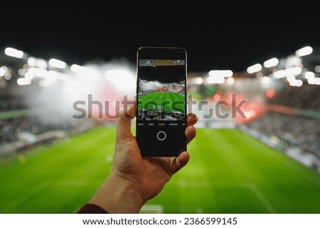 A fan hand is holding a smartphone in a stadium to take pictures of a football match.