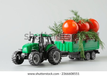 Green toy tractor carries fresh red tomatoes and herbs in a trailer. The concept of agricultural work, harvesting and delivery of crops. Toy world. Photo. Selective focus. Close-up