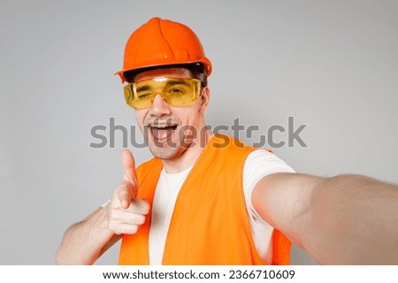 Young employee man in orange vest protective hardhat do selfie on mobile phone point finger camera on you isolated on grey background studio Instruments for renovation apartment. Repair home concept