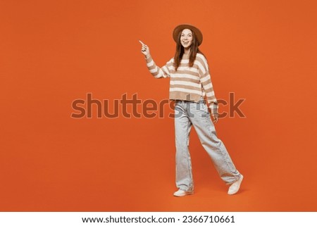 Full body side profile view young woman she wearing striped sweater hat casual clothes walking going waving hand isolated on plain orange red color wall background studio portrait. Lifestyle concept