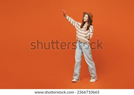 Full body young woman wear striped sweater hat casual clothes doing selfie shot on mobile cell phone post photo on social network show v-sign isolated on plain orange red color wall background studio
