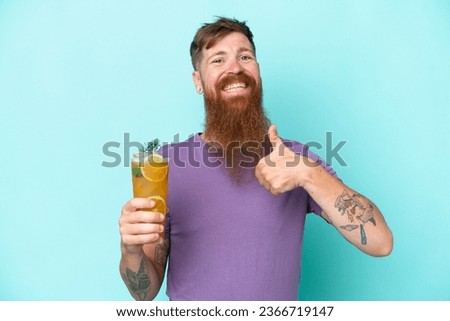 Redhead man with long beard holding a cocktail isolated on blue background with thumbs up because something good has happened