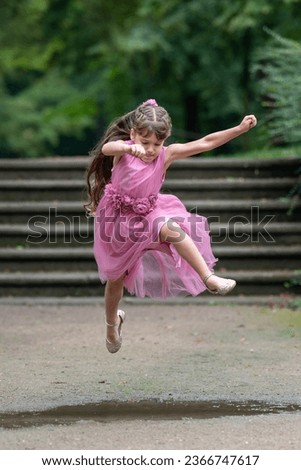 A little girl in a beautiful pink dress with long hair runs quickly and jumps over a puddle. I concentrated and bit my tongue. Outdoor games. Blurred background.