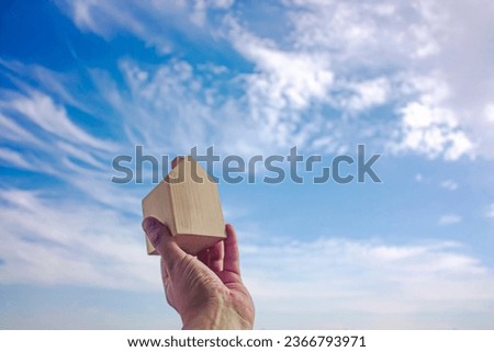 Man holding a small wooden house in palm and sky background. Photograph.