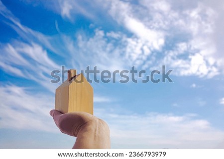 Man holding a small wooden house in palm and sky background. Photograph.