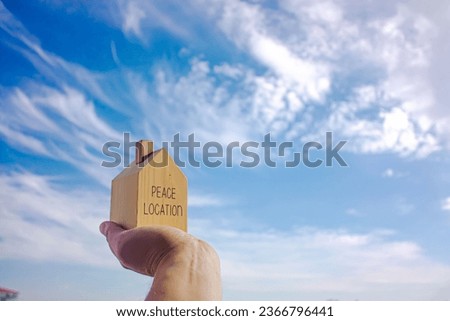 Wooden house with peace location written on it and a human hand holding it. Sky background. Photograph.
