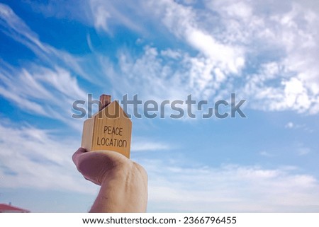 Wooden house with peace location written on it and a human hand holding it. Sky background. Photograph.