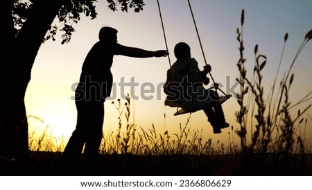 Loving father riding preteen son on swings dark silhouettes in country park against sunset sky. Little boy with father swinging silhouettes at twilight. Father and son play swings in evening field