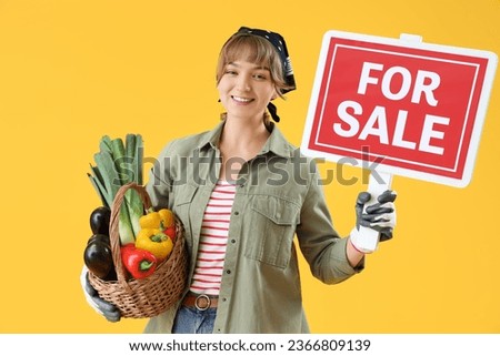 Young female farmer with wicker basket full of different ripe vegetables and FOR SALE sign on yellow background