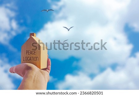Wooden house with 'freedom location' written on it and a human hand holding it. Sky background and birds. Photograph.