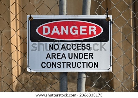 White, black, and red danger no access under construction sign, attached to a rusty chain link fence
