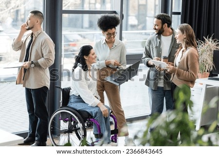 cheerful interracial business people and disabled woman in wheelchair using devices in coworking Royalty-Free Stock Photo #2366843645