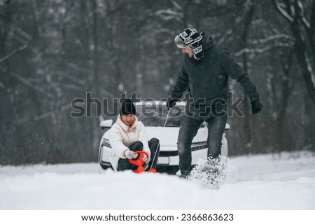 Happy couple are outdoors at winter time. Man takes his wife on a sled. White car behind.