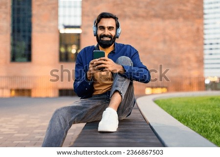 Carefree arabic young man using mobile phone and headphones, relaxing and enjoying online music in urban city area sitting outside, smiling at camera. Musical gadgets, great playlist