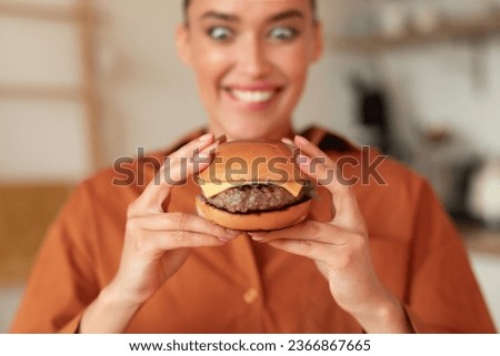 Fast food addiction. Portrait of hungry woman holding burger and looking at it with wide open eyes, enjoying delicious juicy snack, sitting in kitchen, selective focus Royalty-Free Stock Photo #2366867665