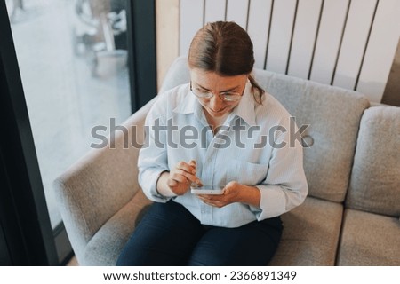 usinesswoman with phone sitting on sofa in office. Freelancer working in coworking space. Female using smartphone to text sms, share photos, communicate with friends, check email, watch videos online