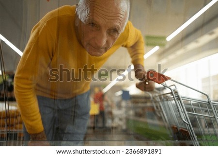 Elderly man shopping in a supermarket with a cart in his hands, looking at the showcase, view through the glass. Grocery store shopping concept Royalty-Free Stock Photo #2366891891