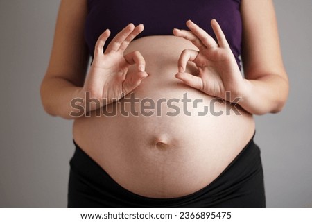 Young pregnant woman standing on gray studio background showing both hands with fingers OK sign, wellness in pregnancy and emotional health concept