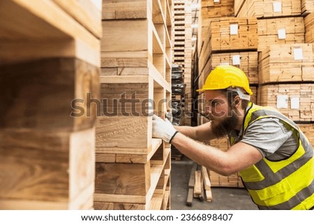 Wood factory worker man checking quality in wood distribution warehouse, male woodman in safety uniform doing quality control, serious factory worker man checking at wood pallets on shelf  Royalty-Free Stock Photo #2366898407