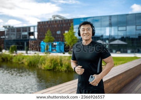 Smiling Asian man jogging outside in the morning, wearing headphones listening to music from a mobile phone.
