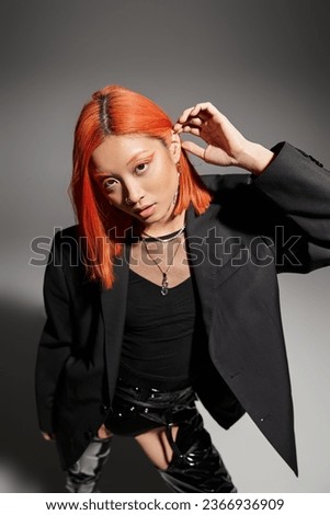 stylish asian woman posing in black blazer and adjusting red hair on grey background, bold style