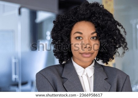 Close up portrait of serious confident business woman, african american woman with curly hair and in business suit looking at camera, female worker inside office at workplace.