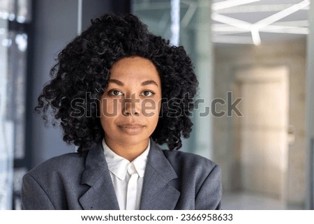 Close up portrait of serious confident business woman, african american woman with curly hair and in business suit looking at camera, female worker inside office at workplace.