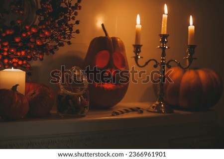 Halloween pumpkin smiling jack lantern and burning candlestick by three candles on fireplace at night home interior. Hallows eve decoration funny glow pumpkin with red berry wreath at dark background