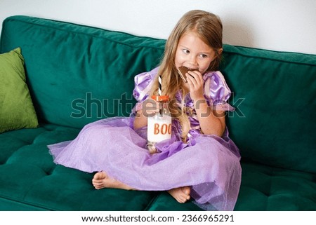 Cute Caucasian little girl is eating cookie in a Halloween costume. Girl in purple dress. Kids and Halloween concept