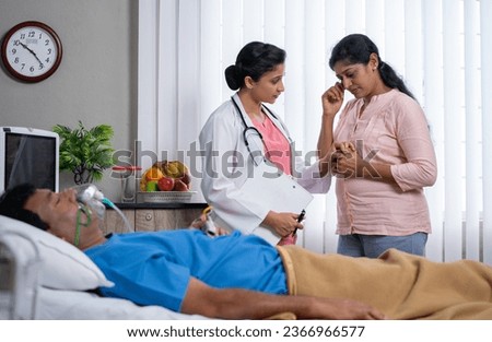 Indian doctor consoling wife of sick patient by giving confidence in front of sleeping husband on bed at hospital ward - concept of encouragement, empowering and medical support.