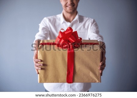 Cropped image of handsome romantic guy smiling while holding a big gift box for his couple, on gray background