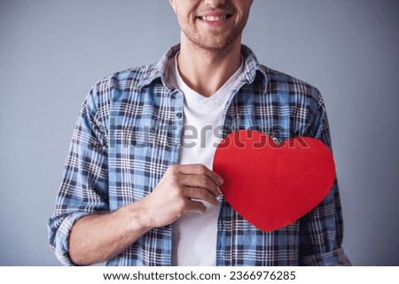 Cropped image of handsome romantic guy smiling while holding a red paper heart, on gray background