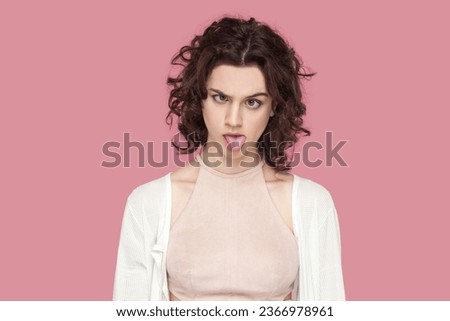 Portrait of crazy fool woman with curly hair wearing casual style outfit having foolish facial expression, grimacing, showing bad manners. Indoor studio shot isolated on pink background. Royalty-Free Stock Photo #2366978961