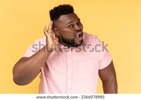I can't hear. Portrait of man wearing pink shirt holding hand near ear, listening attentively with interest private conversation, confidential talk. Indoor studio shot isolated on yellow background. Royalty-Free Stock Photo #2366978985