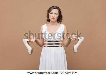 Portrait of sad upset middle aged woman with wavy hair holding elegant shoes on high heels, feels pain on feet, wearing white dress. Indoor studio shot isolated on light brown background. Royalty-Free Stock Photo #2366981991