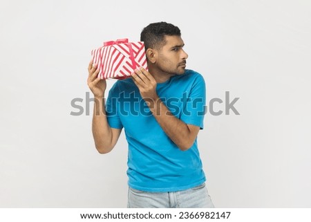Portrait of curious unshaven man wearing blue T- shirt standing with interested facial expression, shaking present box, thinking what inside. Indoor studio shot isolated on gray background.