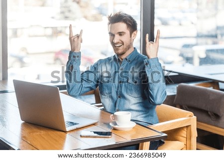 Portrait of crazy excited cool young man freelancer in blue jeans shirt working on laptop, showing rock and roll gesture to computer screen. Indoor shot near big window, cafe background.