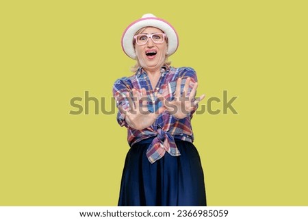 Portrait of mature woman wearing checkered shirt, hat and eyeglasses with puzzled expression, keeps palms in stop gesture, asks to be calm. Indoor studio shot isolated on yellow background.