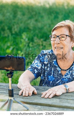 Senior woman recording a podcast and vlogging with smartphone sitting on a park bench